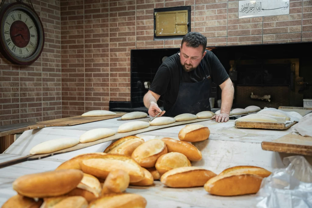 A baker shaping rolls on a floured counter in front of a brick oven in a small independent bakery