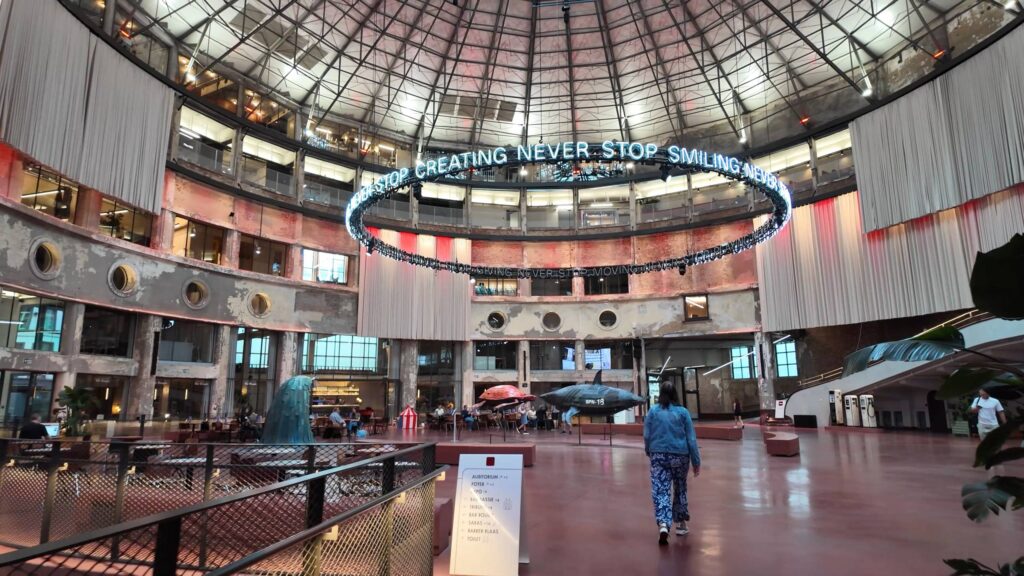Interior of Wintercircus in Ghent: a circular atrium under a glass-and-steel dome, with a ring of blue neon text reading "Never stop creating, never stop smiling" suspended in the centre. Below, people sit at café tables and a woman in a denim jacket walks across the red floor past a large shark sculpture.